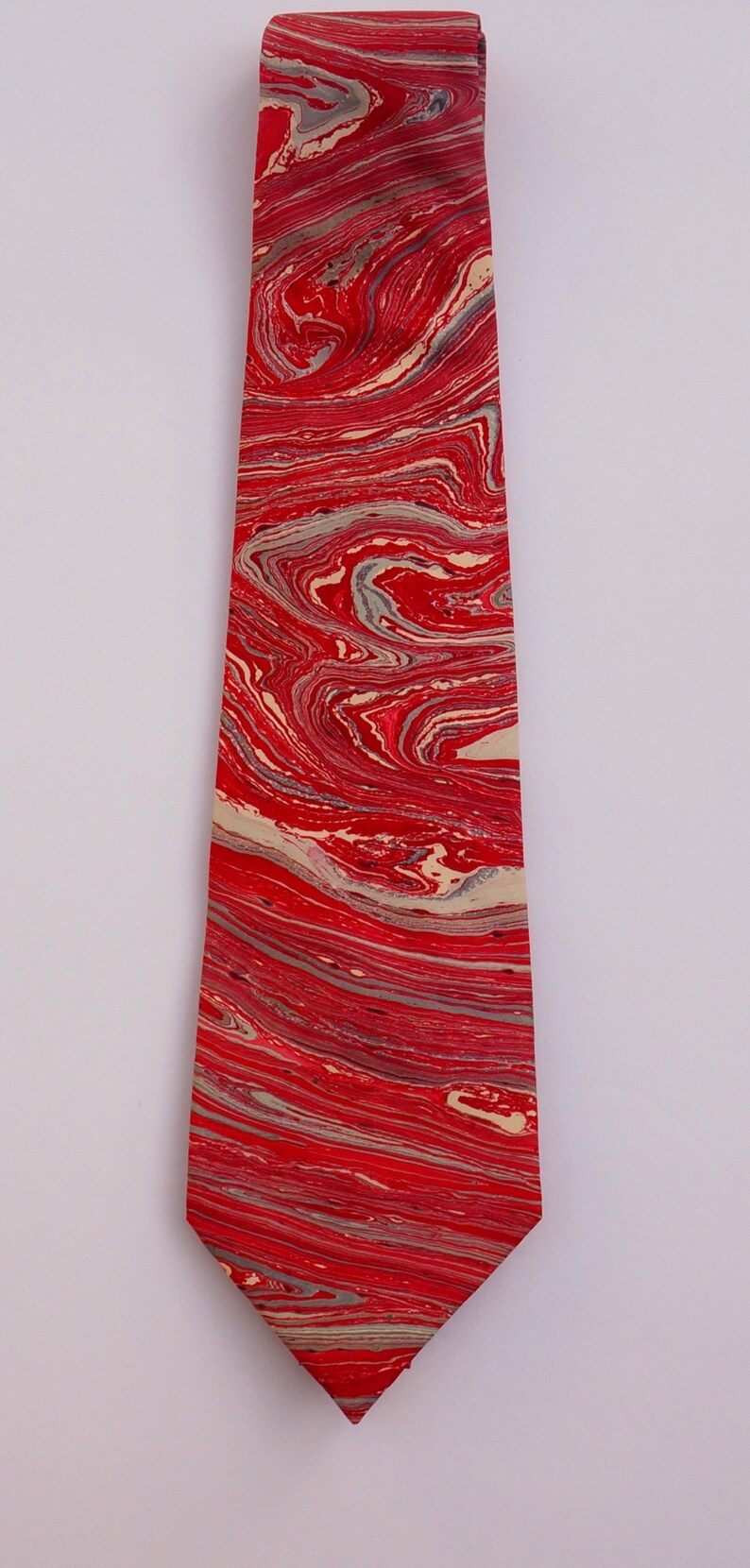 May include: A red and cream tie with a marbled pattern. The tie is made of a smooth fabric and has a pointed tip.