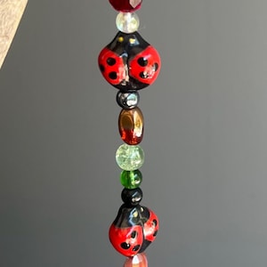 Puede incluir: Adorno de cuentas con cuentas en forma de mariquita rojas y negras, intercaladas con varias cuentas de colores. El adorno está suspendido de un hilo fino, creando una pieza caprichosa y decorativa.