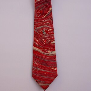 May include: A red and cream tie with a marbled pattern. The tie is made of silk and has a pointed tip.