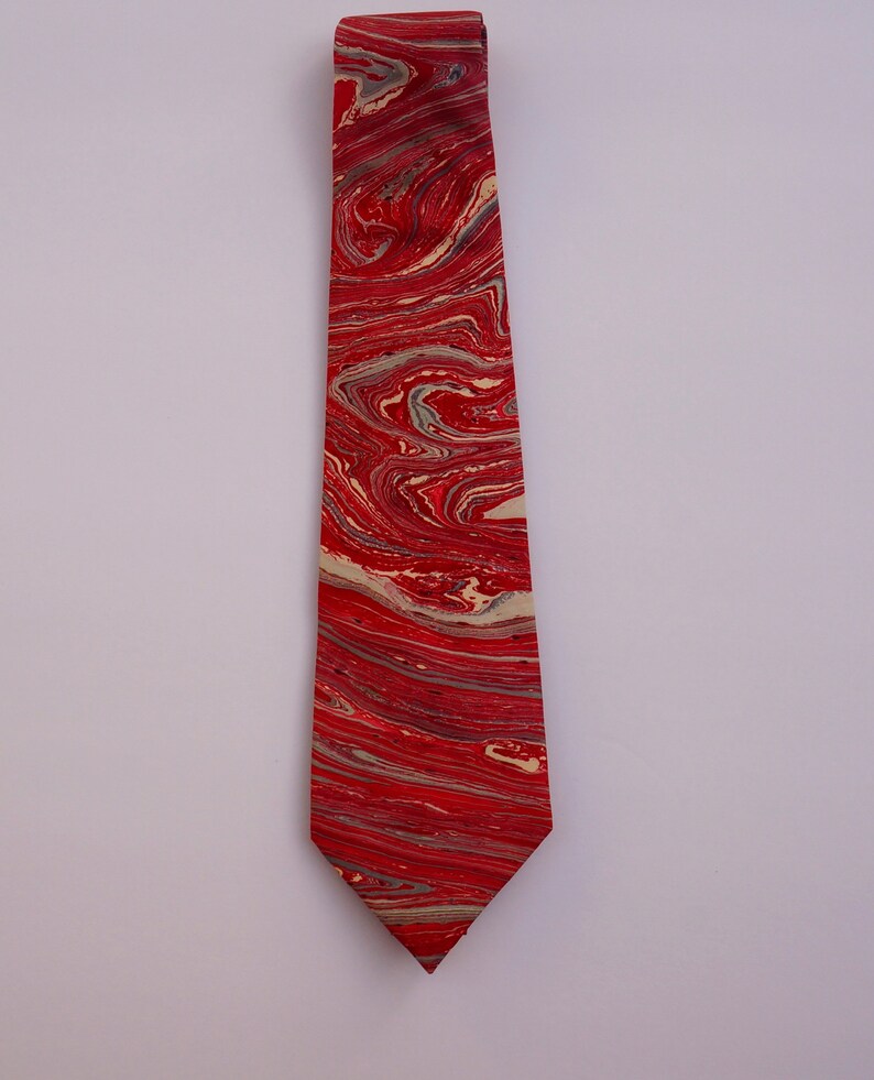 May include: A red and cream tie with a marbled pattern. The tie is made of silk and has a pointed tip.