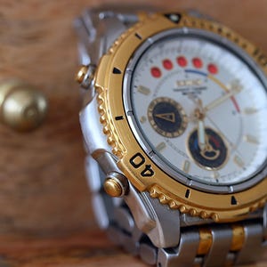 Op de afbeelding: Een zilver- en goudkleurig polshorloge met een witte wijzerplaat en gouden lunette. Het horloge heeft een gedetailleerde wijzerplaat met meerdere sub-wijzerplaten en rode accenten. De band is een combinatie van zilveren en gouden schakels.