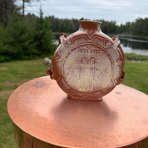 May include: A round, terracotta-colored ceramic canteen with a narrow neck and a flat, circular body. The front features a raised, textured relief design in a lighter, cream-colored glaze, accented by brown leather straps attached to the sides.