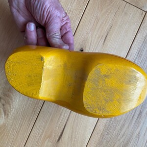 Puede incluir: Un zueco de madera amarillo, visto desde la suela, descansa sobre una superficie de madera de color claro. La pintura amarilla brillante del zueco muestra signos de desgaste. La imagen est&aacute; tomada desde un &aacute;ngulo alto, mostrando la forma y textura del zapato.
