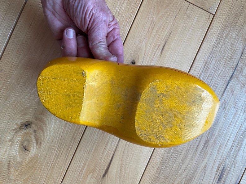 Puede incluir: Un zueco de madera amarillo, visto desde la suela, descansa sobre una superficie de madera de color claro. La pintura amarilla brillante del zueco muestra signos de desgaste. La imagen est&aacute; tomada desde un &aacute;ngulo alto, mostrando la forma y textura del zapato.