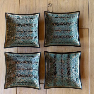 May include: A set of four square dishes with curved sides, featuring a teal and black background with an intricate gold filigree pattern. Each dish has a dark rim and a glossy finish, arranged in a two-by-two grid pattern on a flat surface.