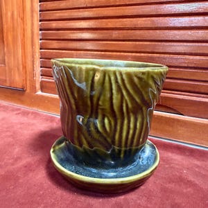 May include: A ceramic planter with a glossy, dark olive green glaze and a textured surface that mimics the appearance of tree bark. The planter includes a matching circular saucer base, featuring a wide opening and a tapered, cylindrical shape.