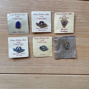 May include: A collection of vintage Camp Archery Association of the United States pins. The pins are on small, aged cards and feature text such as "Junior Bowman," "Silverbow Archer," and "Yeoman." The pins are in various shapes and colors.