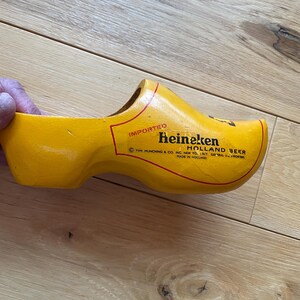 Puede incluir: Un zueco de madera amarillo con el texto "Heineken Holland Beer" en letras rojas. El zueco es un zapato tradicional holand&eacute;s, y el texto indica que es un producto importado. Fabricado en Holanda.