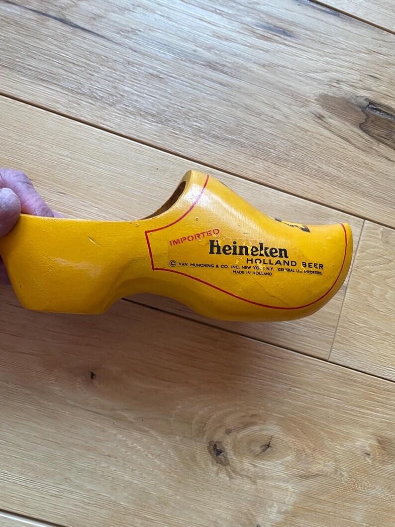 Puede incluir: Un zueco de madera amarillo con el texto "Heineken Holland Beer" en letras rojas. El zueco es un zapato tradicional holand&eacute;s, y el texto indica que es un producto importado. Fabricado en Holanda.