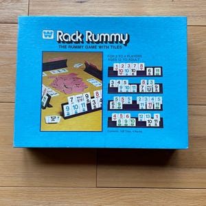 May include: A blue box for the game Rack Rummy, with the game's name and the phrase "THE RUMMY GAME WITH TILES" printed on the front. The box shows game tiles and racks, and the text "FOR 2 TO 4 PLAYERS AGES 12 TO ADULT".