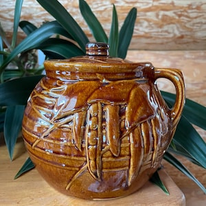 May include: A brown glazed ceramic jar with a lid, featuring a raised design of corn cobs and leaves. The jar has a handle on one side.