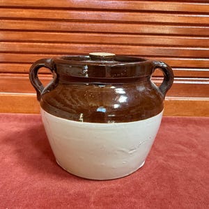 May include: A two-toned ceramic crock with a lid. The upper portion is a glossy brown, while the lower half is a matte cream color. It has two side handles and a small, round lid. This vintage kitchenware is likely used for food storage.