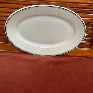 May include: An oval, white ceramic serving platter with a thin black and green border. The platter is displayed on a red surface with a wooden background. This dish is suitable for serving food.