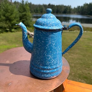 May include: A bright blue enamel coffee pot featuring a white marbled or speckled pattern throughout the exterior. The pot includes a matching lid with a small knob, a curved spout, and a sturdy handle attached to the side of the cylindrical body.
