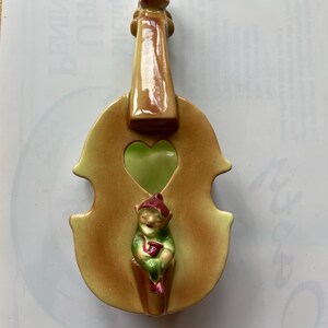 May include: A ceramic violin-shaped wall pocket in shades of brown and yellow, with a green heart-shaped opening. A small elf figure in green and red clothing is seated inside the pocket. The violin has a glossy finish.