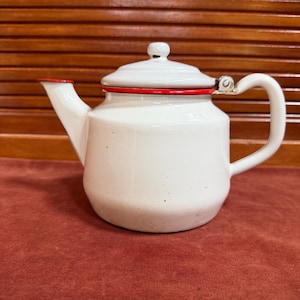 May include: A white enamel teapot featuring a glossy finish, a matching lid with a small knob, a curved handle, and a spout. The rim of the teapot body and the edge of the lid are accented with a thin, vibrant red stripe, providing a distinct contrast.