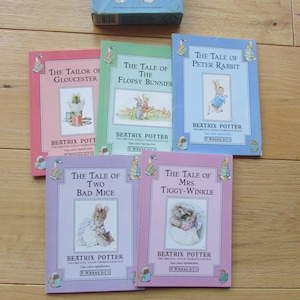 May include: A set of five children's books by Beatrix Potter. The books are all different colors and feature illustrations of the characters from the stories. The titles of the books are "The Tailor of Gloucester", "The Tale of the Flopsy Bunnies", "The Tale of Peter Rabbit", "The Tale of Two Bad Mice", and "The Tale of Mrs. Tiggy-Winkle".