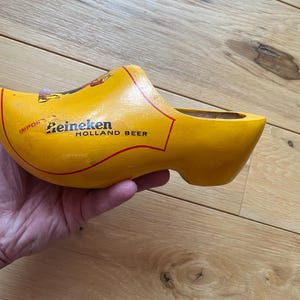 Puede incluir: Un zueco de madera amarillo con el logotipo de Heineken Holland Beer. El zueco es un zapato tradicional holand&eacute;s, con el logotipo impreso en negro y rojo. El zueco est&aacute; sostenido en una mano.