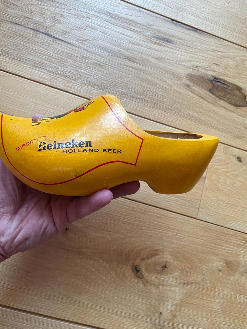 Puede incluir: Un zueco de madera amarillo con el logotipo de Heineken Holland Beer. El zueco es un zapato tradicional holand&eacute;s, con el logotipo impreso en negro y rojo. El zueco est&aacute; sostenido en una mano.