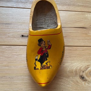 Puede incluir: Un zueco de madera amarillo con una imagen pintada de un personaje de dibujos animados sosteniendo una cerveza. El zueco tiene una franja roja y la palabra "Heineken" est&aacute; impresa en el lateral. Un recuerdo.