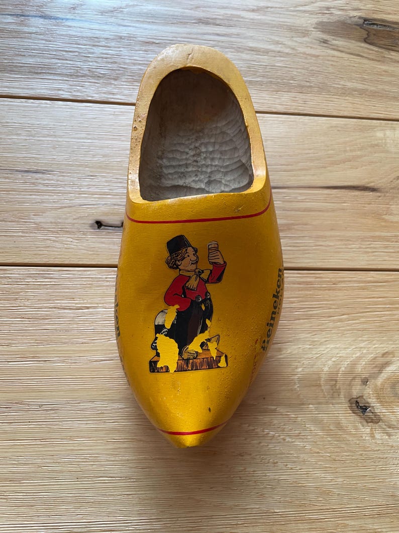 Puede incluir: Un zueco de madera amarillo con una imagen pintada de un personaje de dibujos animados sosteniendo una cerveza. El zueco tiene una franja roja y la palabra "Heineken" est&aacute; impresa en el lateral. Un recuerdo.