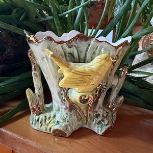 May include: A ceramic planter featuring a sculpted yellow bird perched on a textured tree trunk base. The vessel has a scalloped rim with gold metallic trim, and the exterior is finished in shades of light green and white with gold accents.