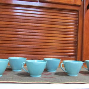 May include: Six teal blue ceramic mugs with rounded handles. The mugs are arranged in a row on a patterned surface.