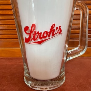 May include: A clear glass pitcher with a red Stroh's logo. The pitcher has a textured base and a handle.