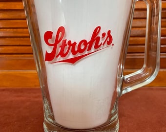 May include: A clear glass pitcher with a red Stroh's logo. The pitcher has a textured base and a handle.