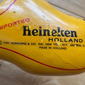 Puede incluir: Un zueco de madera amarillo con la palabra "Heineken" en letras negras. El zueco tiene una raya roja y tambi&eacute;n son visibles las palabras "Imported" y "Holland". El texto "Made in Holland" est&aacute; impreso en el zueco.