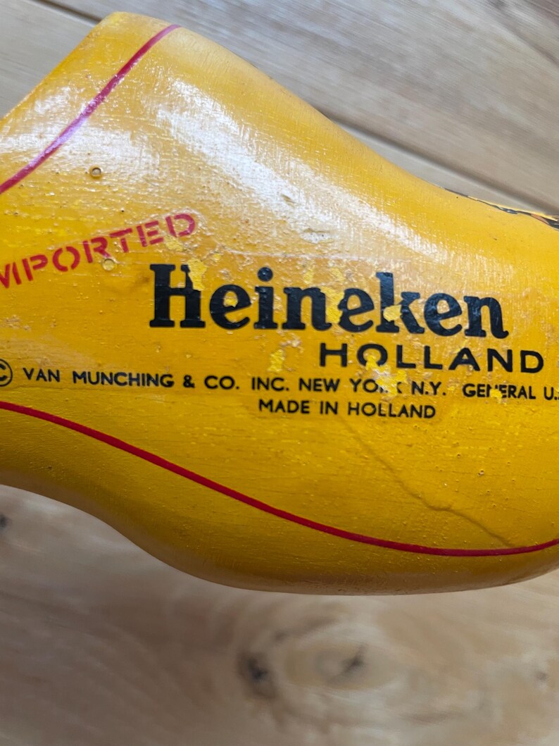 Puede incluir: Un zueco de madera amarillo con la palabra "Heineken" en letras negras. El zueco tiene una raya roja y tambi&eacute;n son visibles las palabras "Imported" y "Holland". El texto "Made in Holland" est&aacute; impreso en el zueco.