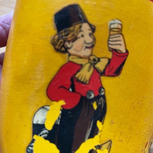 Puede incluir: Un zueco de madera amarillo con una ilustraci&oacute;n pintada de un hombre de dibujos animados sosteniendo una cerveza. El hombre lleva una chaqueta roja, un sombrero de copa negro y una bufanda amarilla. El zueco tiene una raya roja cerca de la parte superior.