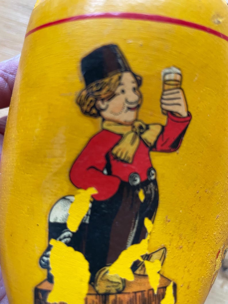 Puede incluir: Un zueco de madera amarillo con una ilustraci&oacute;n pintada de un hombre de dibujos animados sosteniendo una cerveza. El hombre lleva una chaqueta roja, un sombrero de copa negro y una bufanda amarilla. El zueco tiene una raya roja cerca de la parte superior.