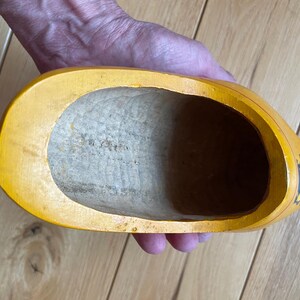 Puede incluir: Un zueco de madera amarillo, sostenido en una mano, con un interior ahuecado. El zueco tiene una punta afilada y un dise&ntilde;o pintado en el lateral. El interior es de color madera clara.