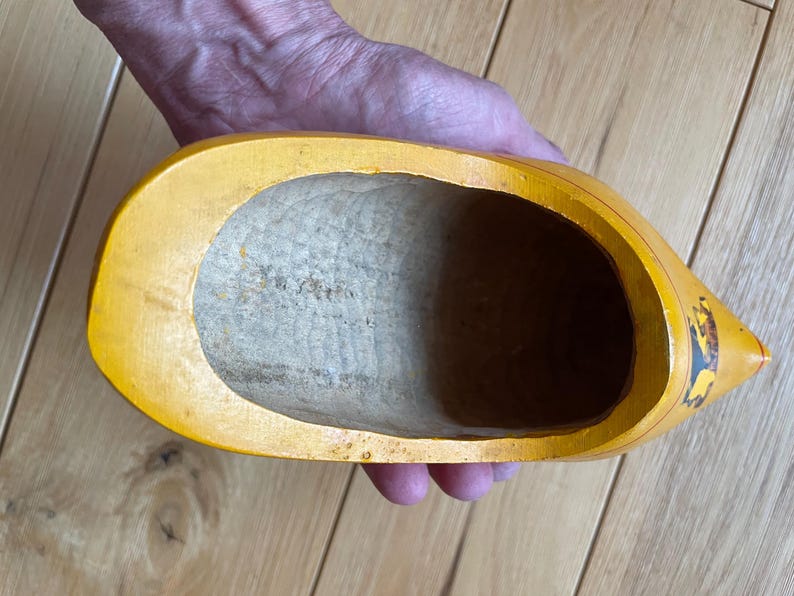 Puede incluir: Un zueco de madera amarillo, sostenido en una mano, con un interior ahuecado. El zueco tiene una punta afilada y un dise&ntilde;o pintado en el lateral. El interior es de color madera clara.