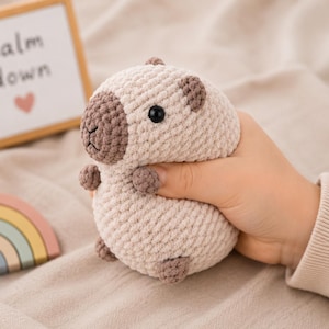 Peut inclure: Un petit jouet en peluche capybara crocheté, de forme ronde, fait de fil beige clair avec un museau, des oreilles et des pattes marron foncé. Le jouet présente des yeux en plastique noir et une bouche cousue. Un panneau indique calm down.