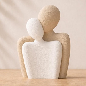 Könnte beinhalten: Abstrakte Skulptur in Beige und Weiß, die zwei sich umarmende Figuren darstellt. Die strukturierte Skulptur steht auf einer Holzoberfläche. Die Figuren sind mit glatten, abgerundeten Formen stilisiert, was ein Gefühl der Einheit erzeugt.