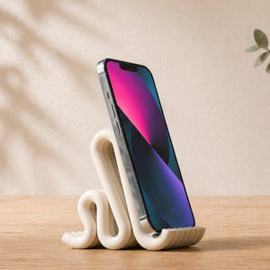 Könnte beinhalten: Ein weißer, wellenförmiger Telefonständer hält ein Smartphone mit farbenfrohem Bildschirm. Der Ständer besteht aus einem glatten, geschwungenen Material. Eine dekorative Vase mit Grün steht im Hintergrund auf einer Holzoberfläche.
