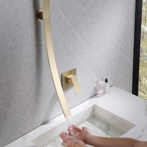 Può includere: Un rubinetto dorato curvo con maniglia quadrata e montaggio a parete. L'acqua scorre dal rubinetto in un lavandino bianco. Il rubinetto è montato su una parete grigia con motivo marmoreo. L'immagine mostra un moderno accessorio per il bagno.