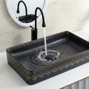 May include: A rectangular black ceramic bathroom sink with a decorative gold pattern around the edges. A black faucet is positioned above the sink, with water flowing into the basin. A peach-colored soap dispenser and a small green plant are on the counter.