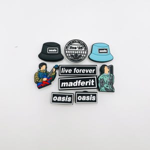 May include: A collection of nine rubberised decorative charms featuring Oasis band themes. Items include a black bucket hat, a light blue bucket hat, a circular Live '25 logo, two rectangular Oasis logos, two rectangular text bars reading live forever and madferit, and two figures of musicians.