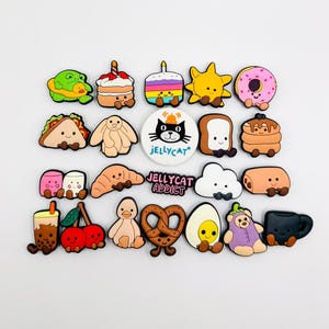 May include: A selection of colourful, cartoon-style charms depicting food and characters. Includes a frog, cake, taco, croissant, and a black cat with the text "Jellycat". The charms are designed for decorative purposes.