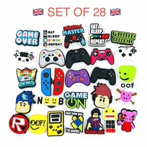 May include: A collection of 28 gaming-themed charms in various colours. The charms include game controllers, character faces, and phrases such as "Game Over," "Eat Sleep Repeat," and "Roblox Master." The charms are in a variety of colours, including black, blue, red, and green.