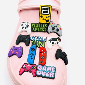May include: A light pink shoe decorated with vibrant video game charms. The charms feature game controllers in black, white, grey, purple, and pink, plus the phrases "GAME ON" and "GAME OVER". A yellow handheld console charm is also visible, creating a playful aesthetic.