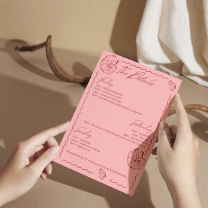 以下が含まれることがあります： 「The Details」のテキストと週末イベントのスケジュールが記載されたピンク色の招待状。カードには貝殻のイラストと波型のボーダーが描かれています。テキストには日付、時間、アクティビティが含まれています。