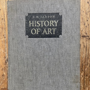 May include: A hardcover book titled "History of Art" by H.W. Janson. The book has a gray fabric cover with a dark gray rectangular label featuring gold lettering. The book is closed and resting on a wooden surface.