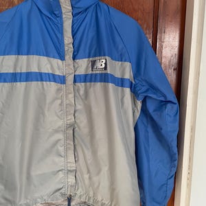 May include: A blue and gray windbreaker jacket with a New Balance logo on the front. The jacket has a zipper closure and a drawstring at the bottom.