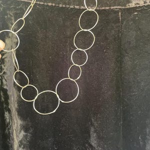 May include: A black velvet skirt featuring a high waistband and a long, straight silhouette. A silver-toned necklace consisting of multiple interlocking circular metal rings of varying sizes is draped over the front of the dark fabric.