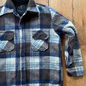 May include: A vintage plaid shirt in shades of blue, brown, and white. The shirt features two chest pockets with button closures and a classic collar. The fabric appears to be a thick, warm material, ideal for cooler weather.