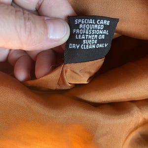 May include: Close-up of a brown leather jacket's care label. The label reads "Special care required professional leather or suede dry clean only."